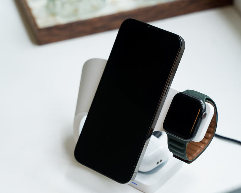 White SIMPLI MagSafe charger with an iPhone and an Apple Watch on a charging dock, featuring an elegant design and placed on a white surface with a wooden background for a minimalist aesthetic.