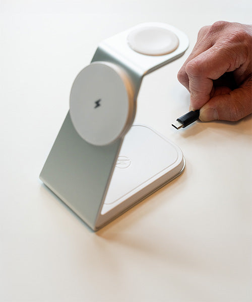 Close-up of a hand plugging a USB-C cable into Magnitis 3-in-1 MagSafe charging station with dual wireless charging pads for Apple devices, highlighted by a minimalist white design on a clean surface.