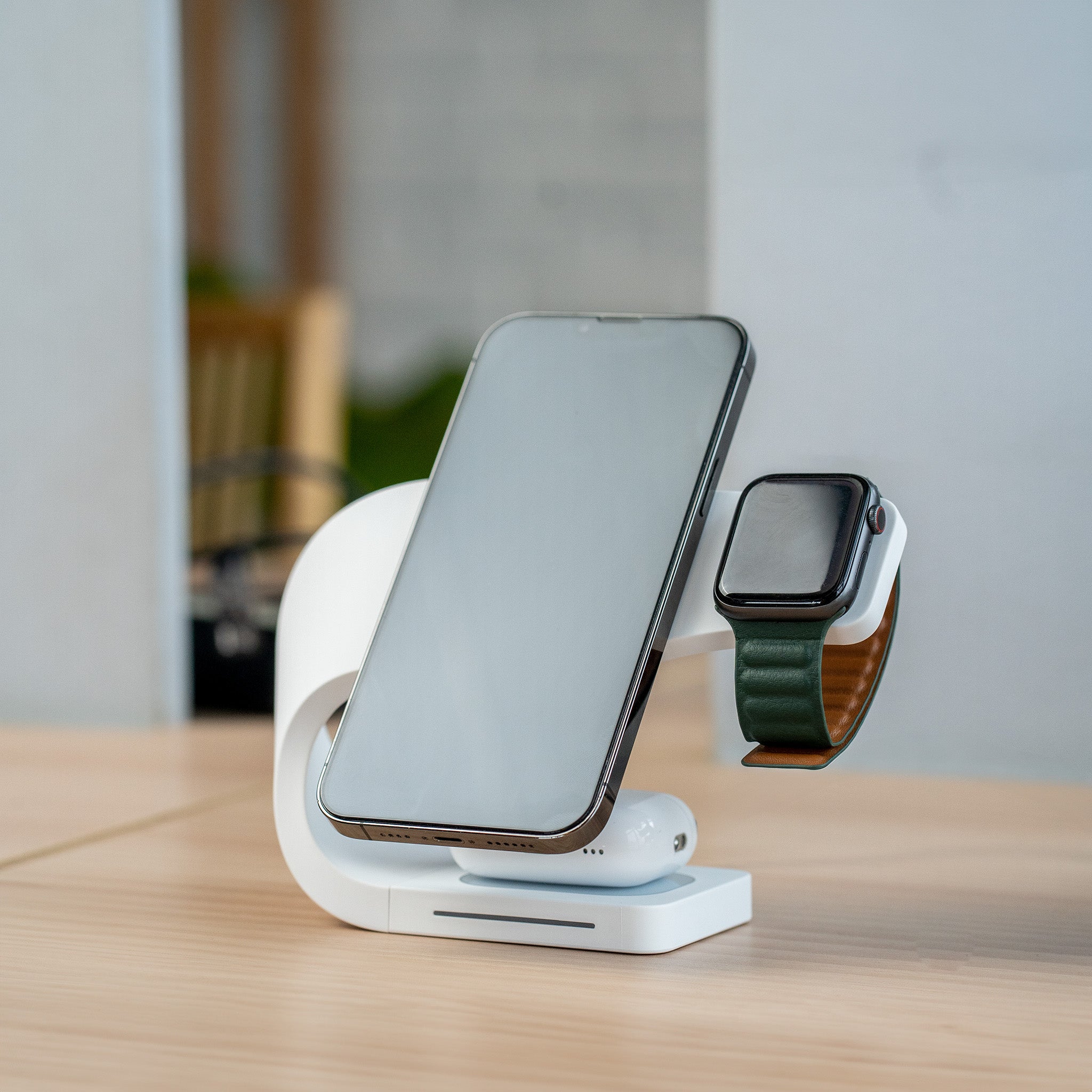 An iPhone and Apple Watch are charging on a Simpli 3-in-1 Wireless Charging Station in Ivory White, placed on a wooden desk. The charger's modern design holds the iPhone vertically and the watch on a side arm, while AirPods rest on the base.